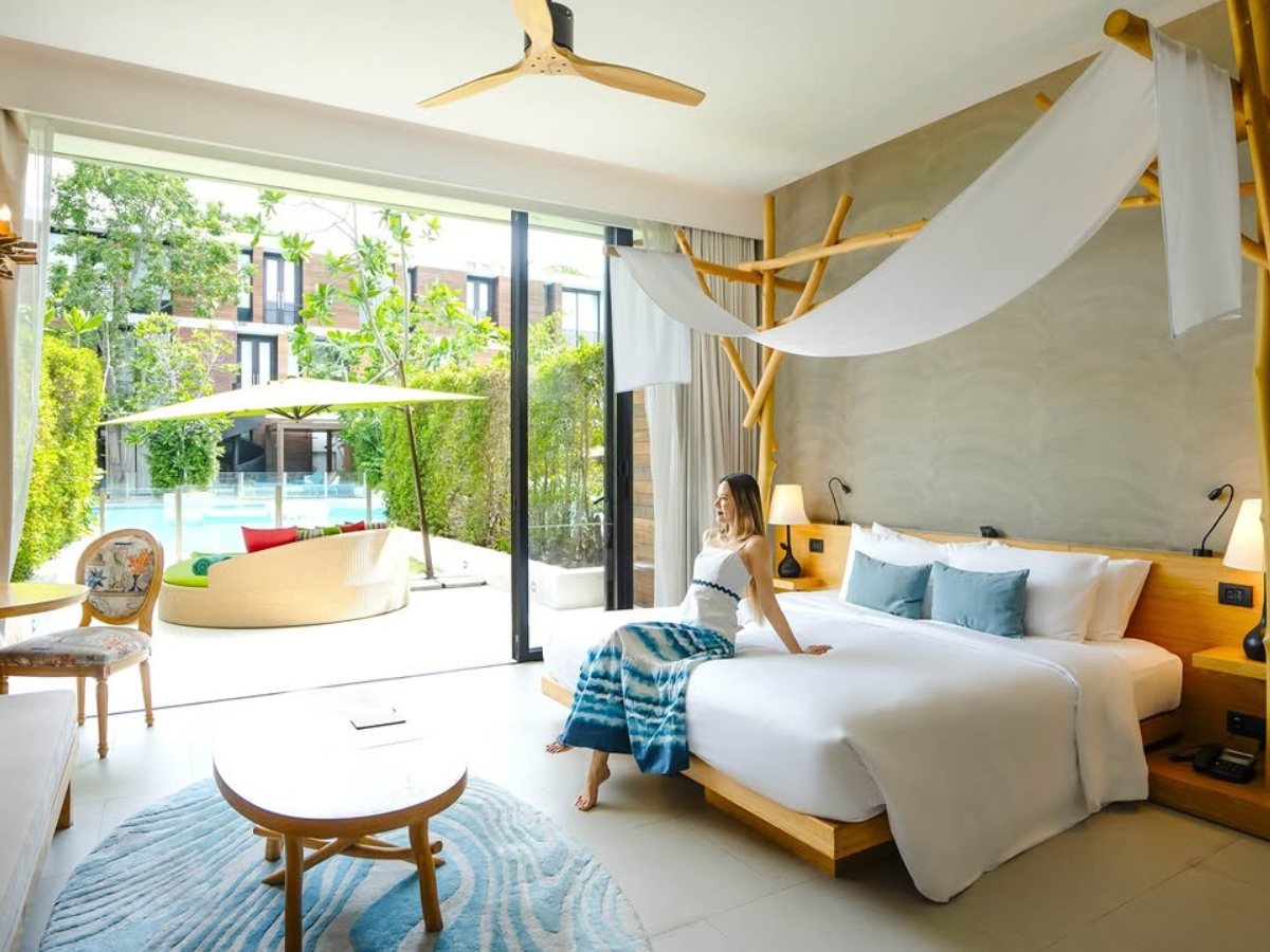 Modern hotel room with a large bed, wooden accents, and a canopy. A woman sits on the bed, gazing outside at a pool through sliding glass doors.