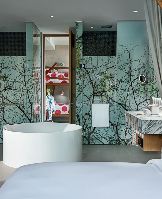 A modern bathroom features a large white circular bathtub, tree-themed wallpaper, and a marble sink. Bunk beds with red polka-dot bedding are visible in the background.