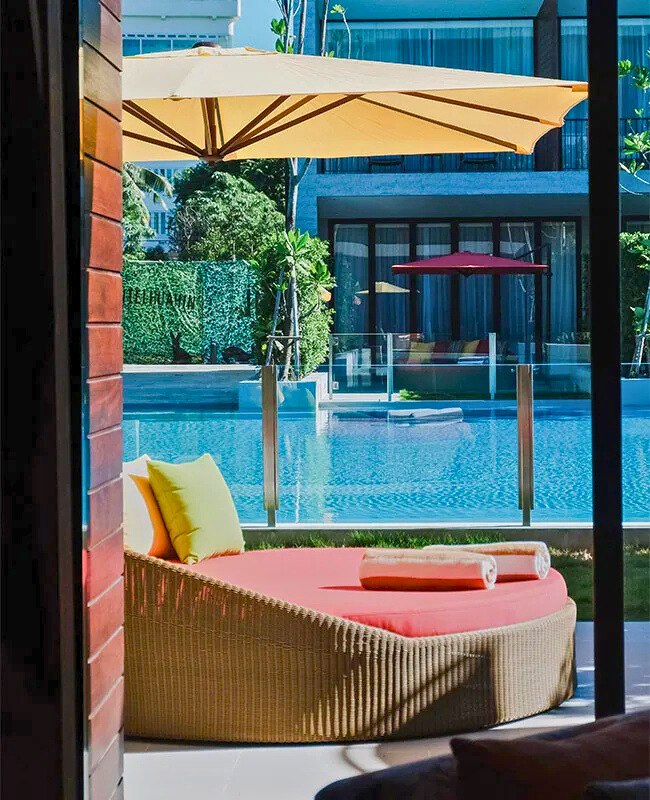 Sunny poolside scene with a round wicker daybed, red cushion, yellow pillows, and rolled towels. Large umbrella and modern building in the background.