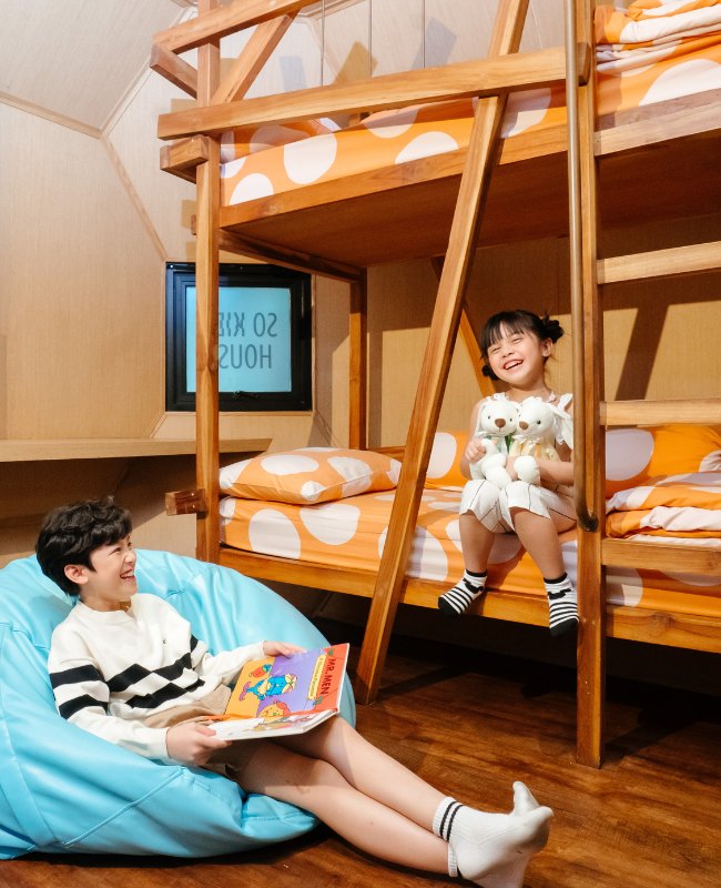 A girl sits on a wooden bunk bed holding toys, smiling at a boy on a bean bag reading a book. The room is cozy with orange polka-dot bedding.