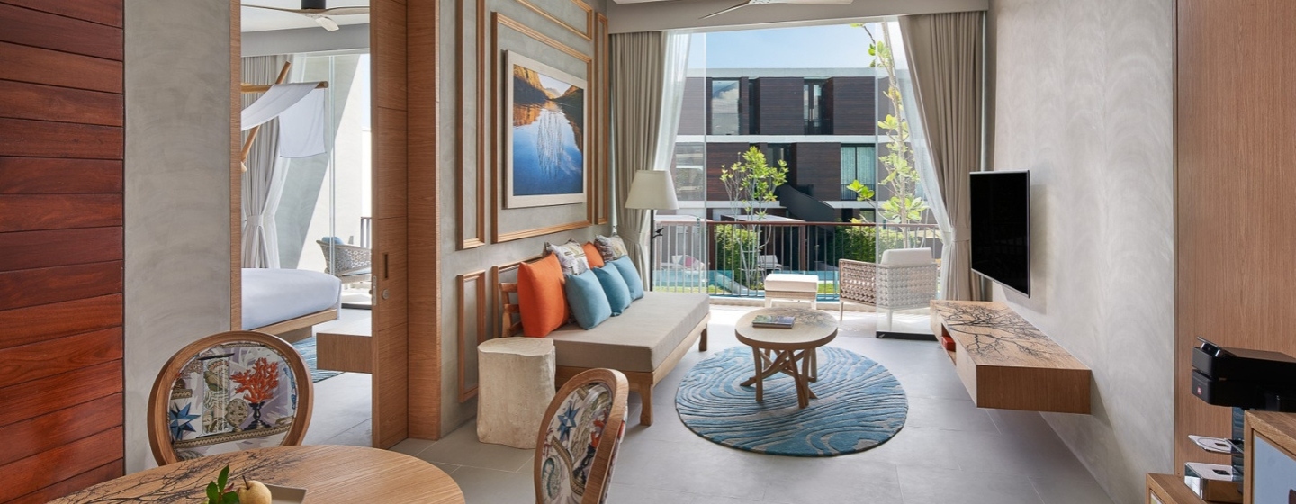 Stylish hotel room with modern decor, featuring a cozy seating area with colorful pillows, a TV, and a balcony view of a green courtyard. Airy and inviting.