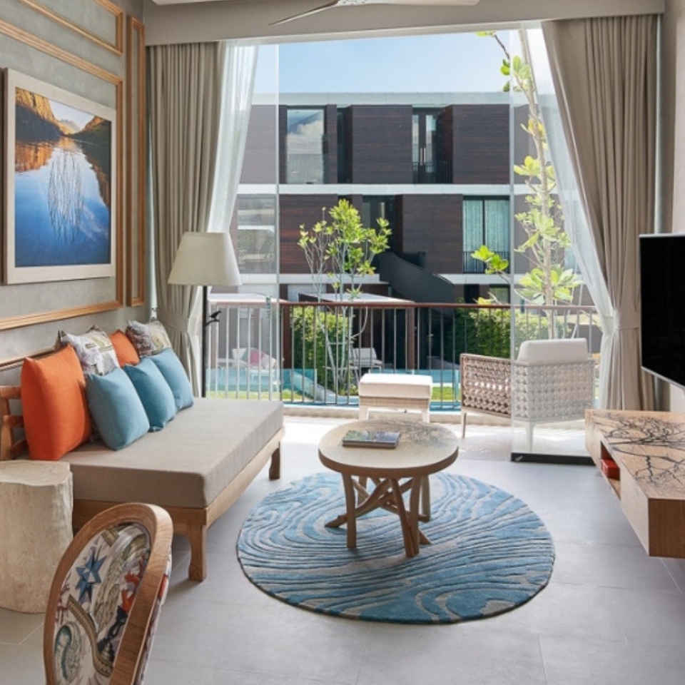 Stylish hotel room with modern decor, featuring a cozy seating area with colorful pillows, a TV, and a balcony view of a green courtyard. Airy and inviting.
