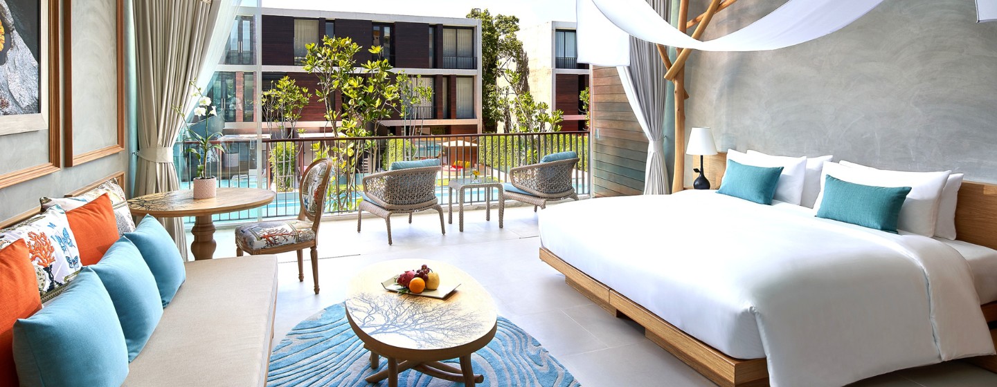 Modern hotel room with large bed, turquoise pillows, wooden accents, and blue rug. Open balcony shows pool view and greenery, creating a relaxing vibe.