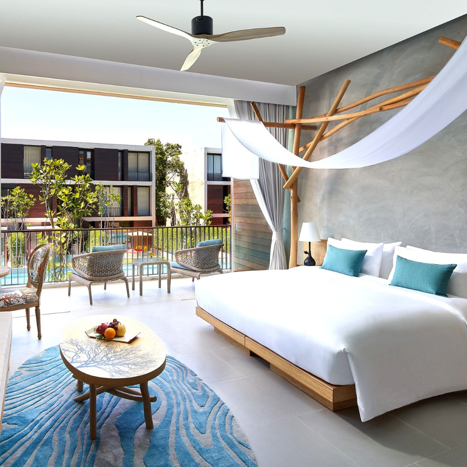 Modern hotel room with large bed, turquoise pillows, wooden accents, and blue rug. Open balcony shows pool view and greenery, creating a relaxing vibe.