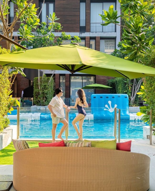 A man and woman chat by a sunny pool, surrounded by green umbrellas and tropical plants. The setting is relaxed and inviting, with colorful seating.
