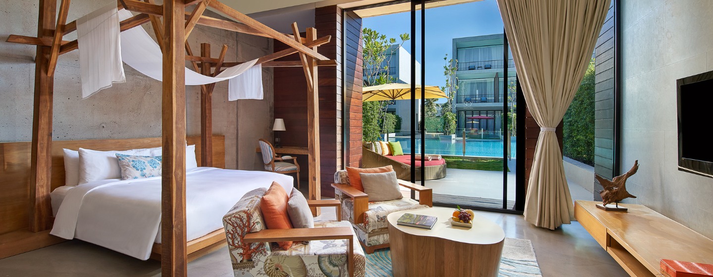 Modern hotel room with a wooden canopy bed, patterned armchairs, and a textured rug. Glass doors open to a pool area with yellow umbrellas, creating a serene ambiance.