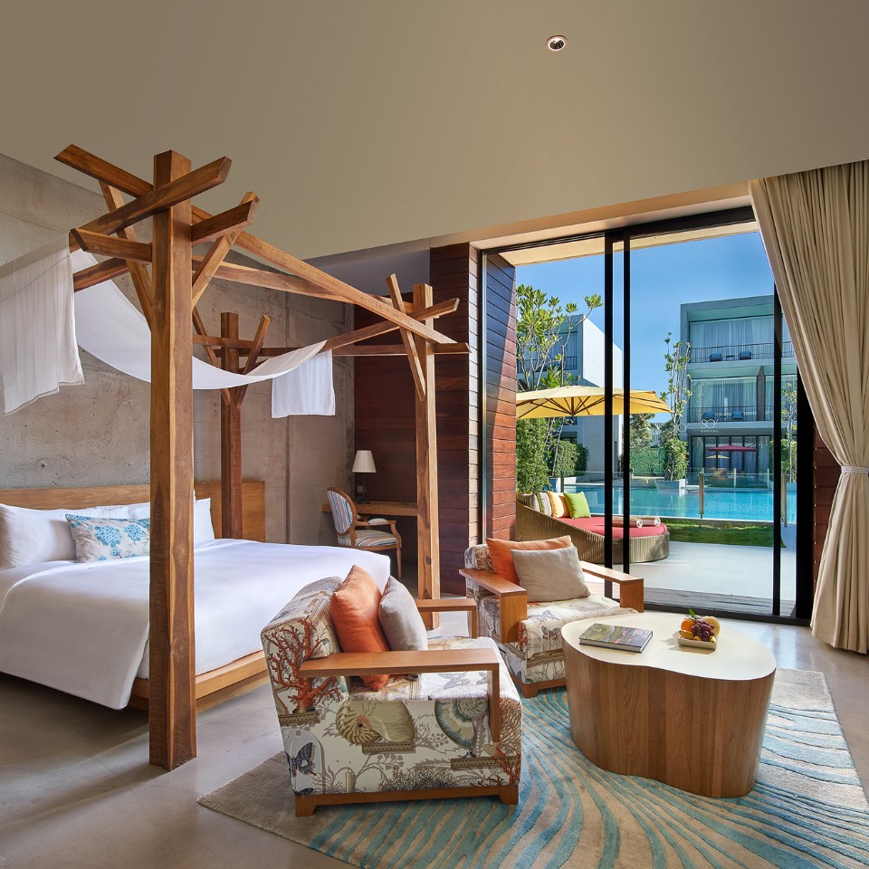 Modern hotel room with a wooden canopy bed, patterned armchairs, and a textured rug. Glass doors open to a pool area with yellow umbrellas, creating a serene ambiance.