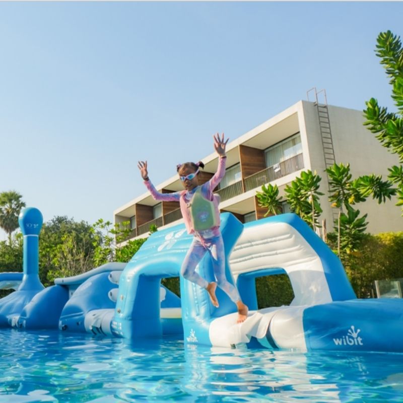 A child in a black and red swimsuit with a life vest joyfully climbs a blue inflatable in a pool. Lush greenery and a modern building backdrop.