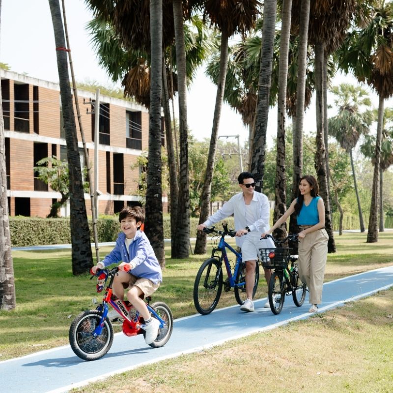 A family enjoying a fun and active outdoor experience, riding bicycles together on the dedicated blue cycling track shaded by palm trees at SO/ Sofitel Hua Hin.