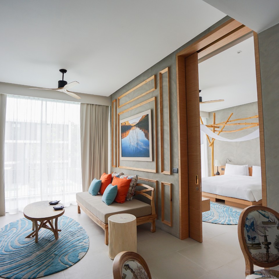 Modern hotel room with natural light, featuring a sofa with orange and teal cushions, a round table, and a bed with white linens. Cozy and inviting.