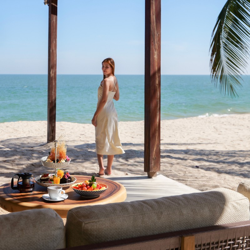 Woman in white dress stands on a sandy beach under a wooden canopy, with a tropical ocean view. Table in foreground with fruit and drinks.