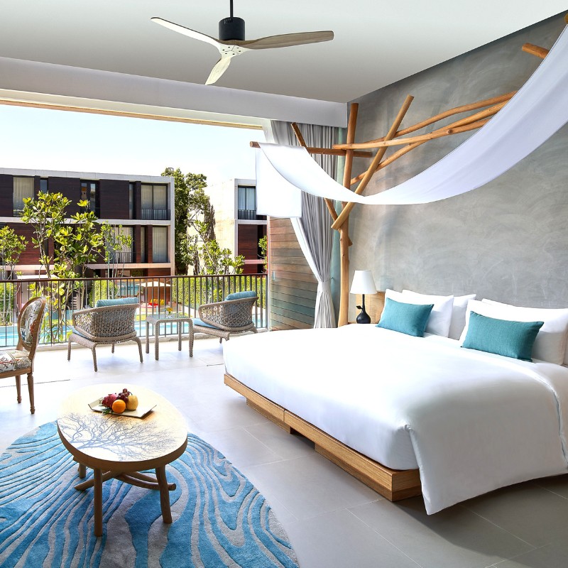 Modern hotel room featuring a large bed with teal pillows, a stylish wood accent wall, open balcony with wicker chairs, and pool view, conveying calm.