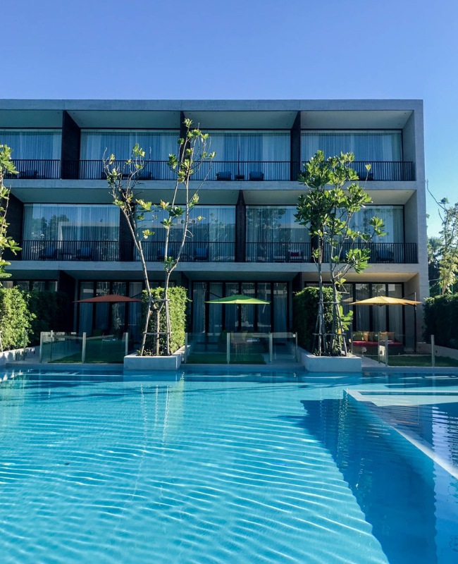 Modern hotel with glass balconies overlooking a sparkling blue pool, surrounded by lush greenery and vibrant umbrellas. The scene is serene and inviting.