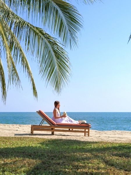A woman in a pink dress relaxes on a wooden lounge chair by the beach under palm leaves, holding a drink. The scene is peaceful and sunny.