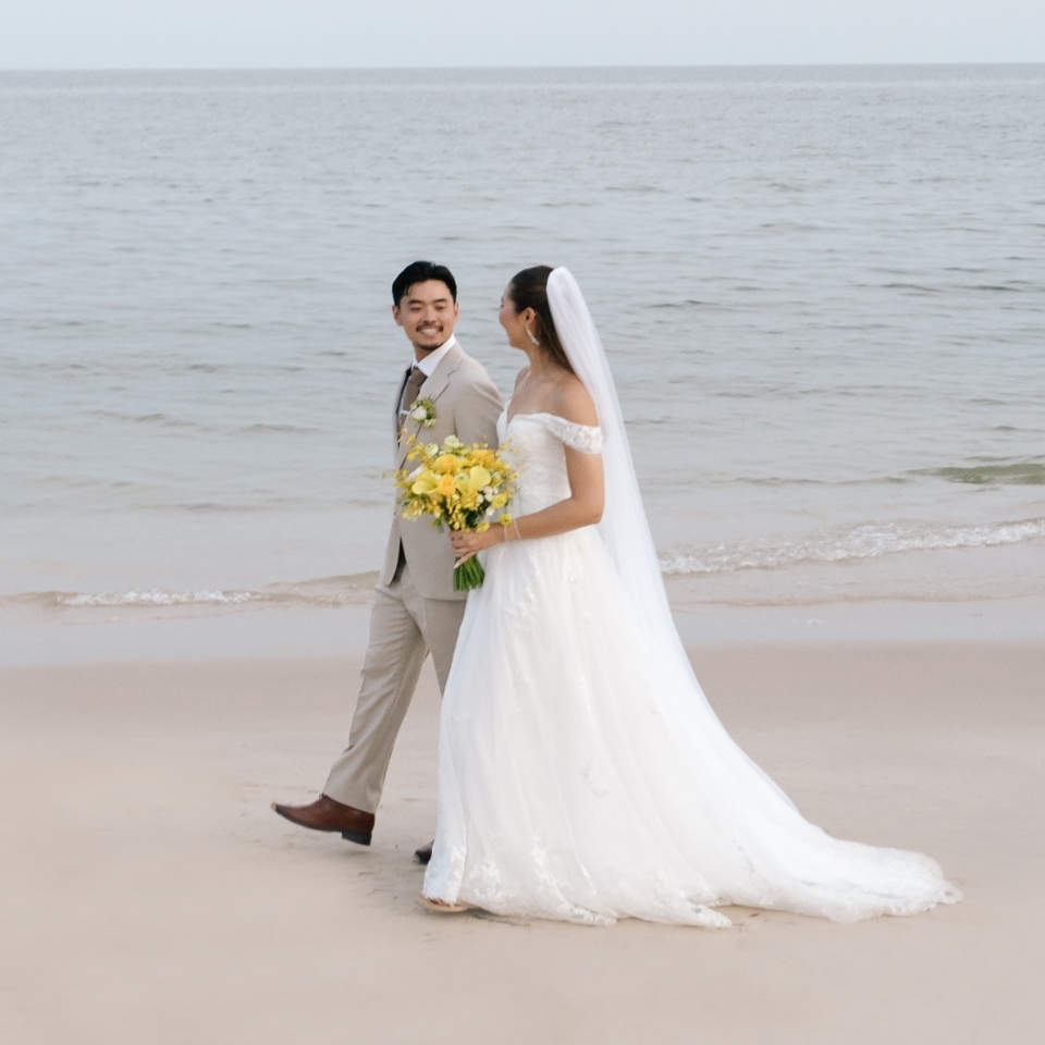 A bride in a flowing white gown holds yellow flowers, walking hand-in-hand with the groom in a light suit along a serene beach by the ocean. Romantic ambiance.