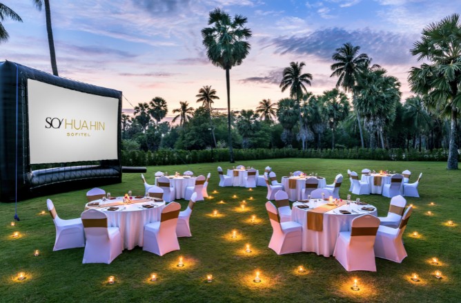 http://Outdoor%20event%20setup%20with%20round%20tables,%20white%20chairs,%20and%20gold%20accents%20surrounded%20by%20candlelit%20lanterns%20on%20grass.%20Large%20screen%20displays%20SO/%20Hua%20Hin%20Sofitel%20amidst%20palm%20trees%20at%20dusk.
