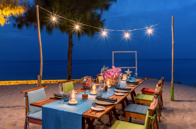 http://Oceanfront%20dining%20setup%20under%20string%20lights%20on%20a%20sandy%20beach%20at%20night.%20A%20long%20table%20with%20candles,%20plates,%20and%20vibrant%20flowers%20creates%20a%20cozy,%20inviting%20atmosphere.