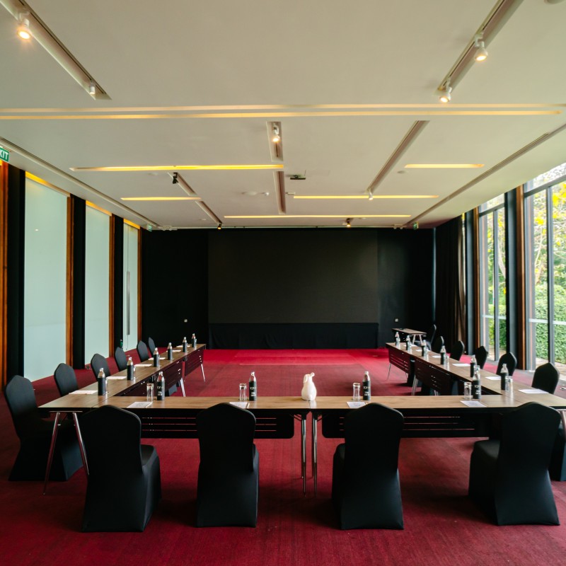 A conference room with red carpet, U-shaped table layout, black chairs. Bottles and notepads neatly placed, large windows to the right, projector screen ahead.