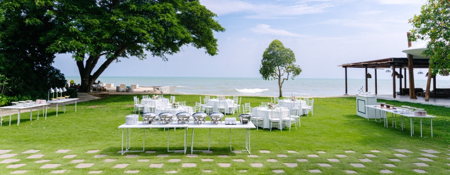 Outdoor buffet setup on a lush green lawn by the sea. Tables with white linens and silver chafing dishes, framed by trees under a clear blue sky.