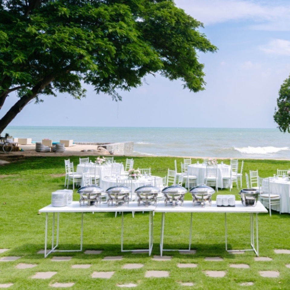 Outdoor buffet setup on a lush green lawn by the sea. Tables with white linens and silver chafing dishes, framed by trees under a clear blue sky.