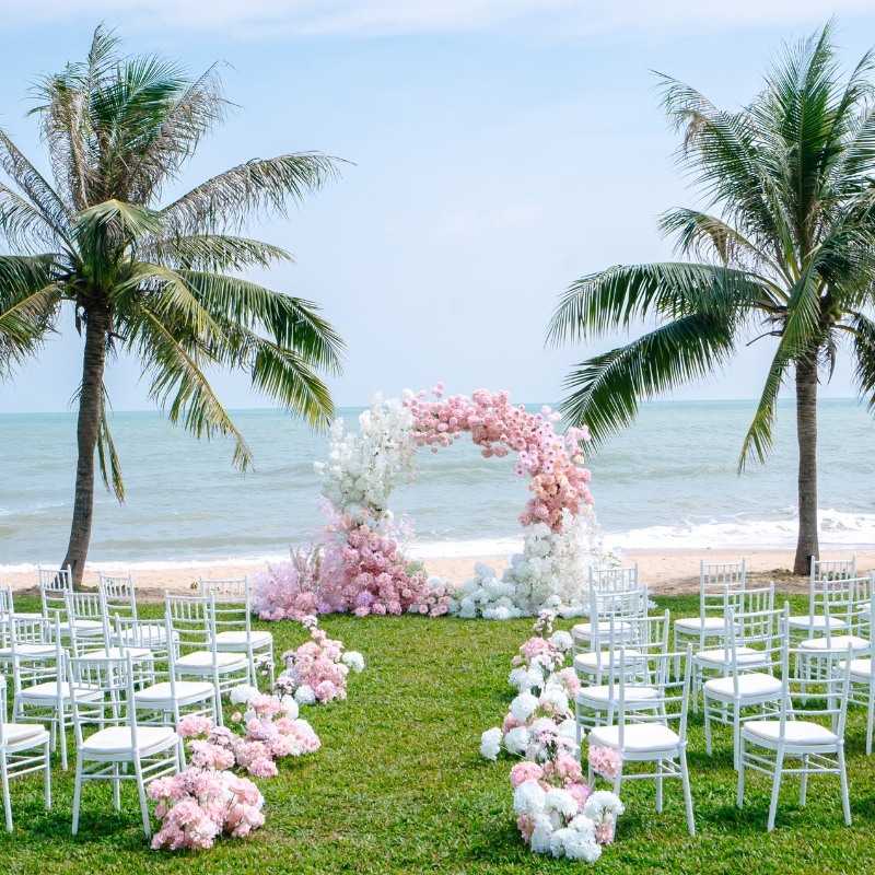Beach wedding setup with a floral arch of pink and white flowers between two palm trees, surrounded by white chairs on green grass, overlooking the ocean.