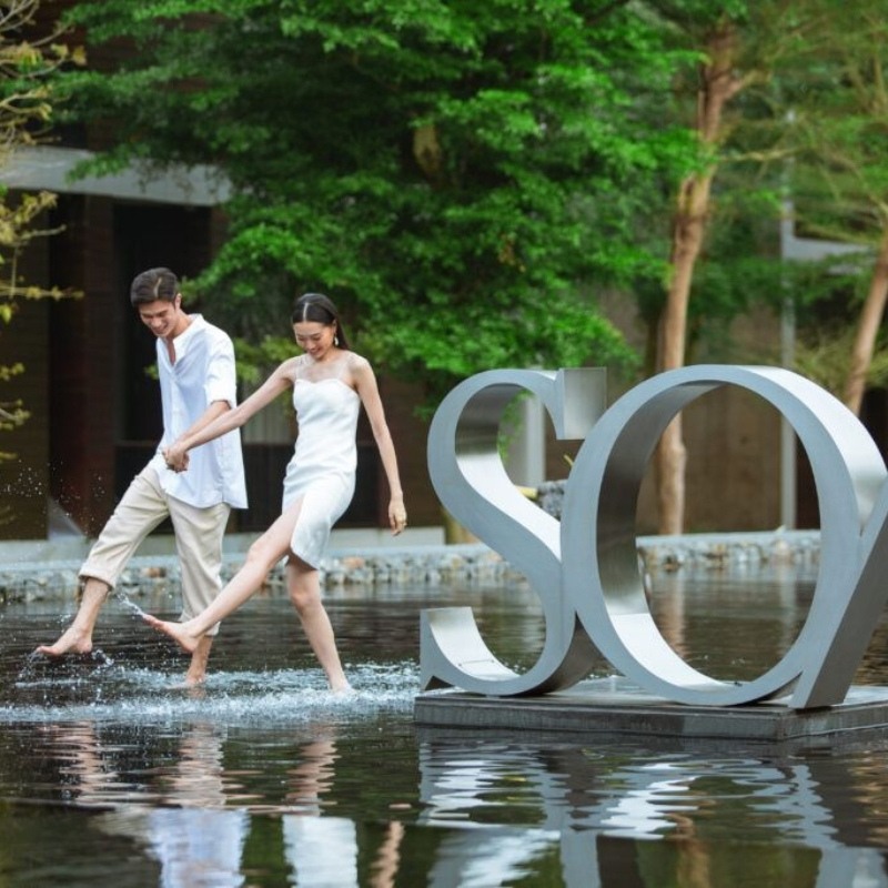 A couple joyfully walks hand in hand through a shallow reflective pool, barefoot and smiling, near a large "SO/" sculpture. The scene is serene and playful.