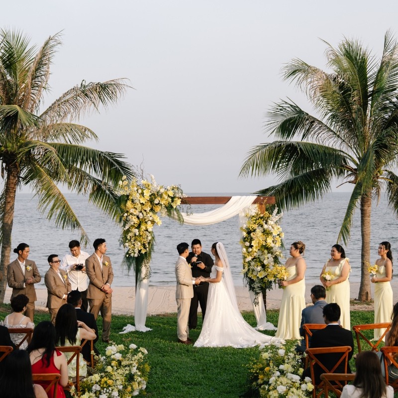A couple stands at an outdoor wedding ceremony by the ocean, framed by palm trees and floral arches. Guests seated on wooden chairs watch.