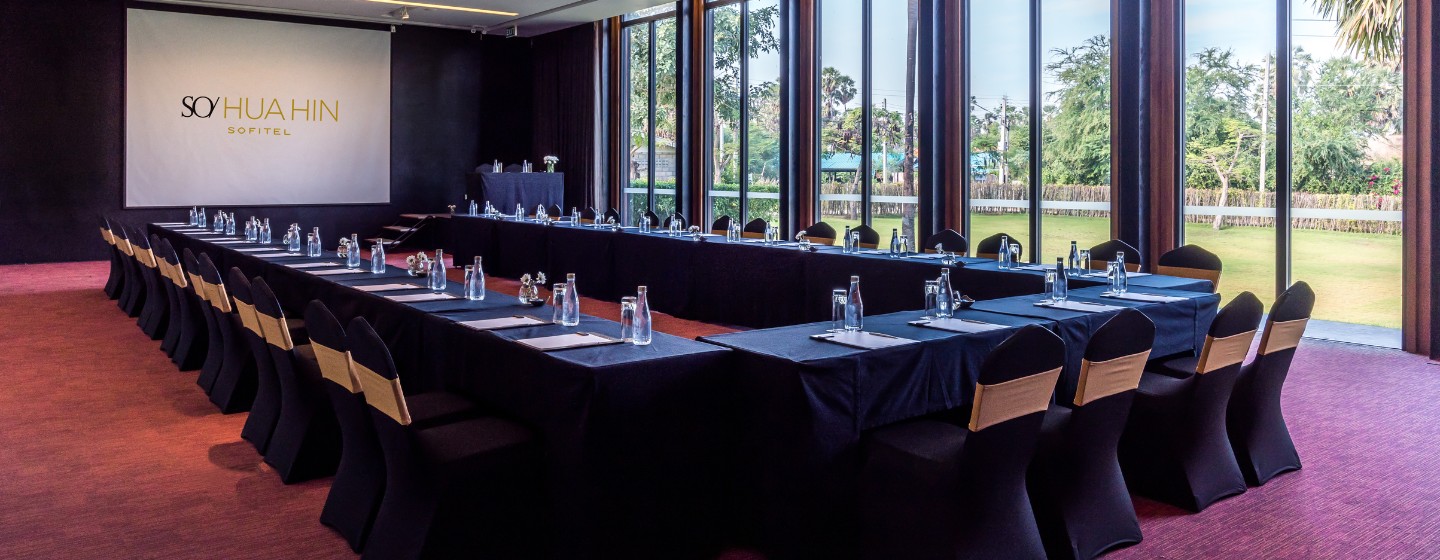 A conference room with a U-shaped table setup, black tablecloths and chairs, water bottles, and notepads. Large windows offer views of greenery outside.
