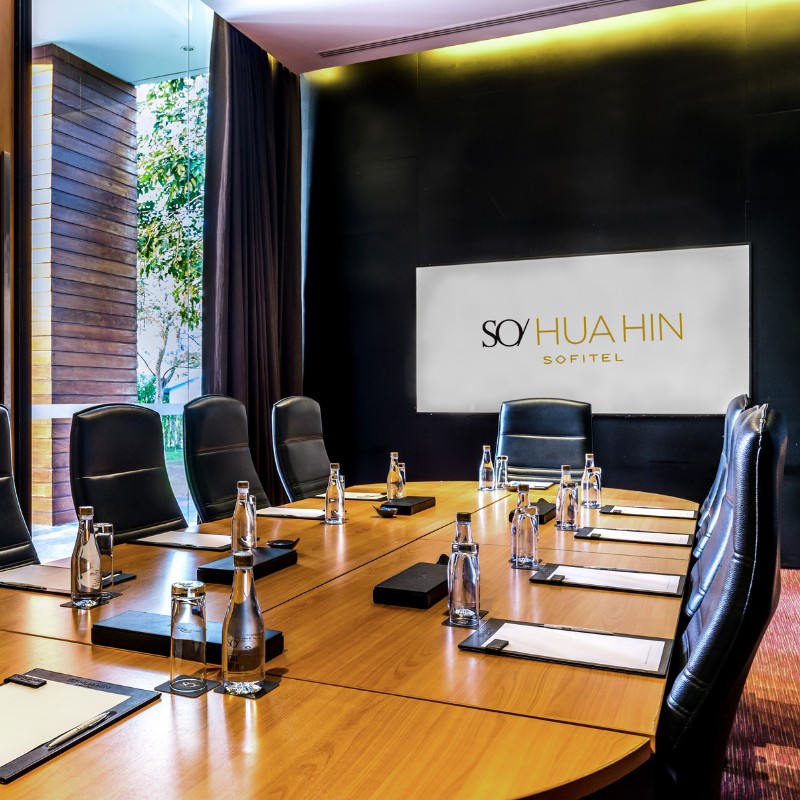 A modern conference room with a wooden table, black leather chairs, and water bottles. A large window lets in light, creating a professional ambiance.