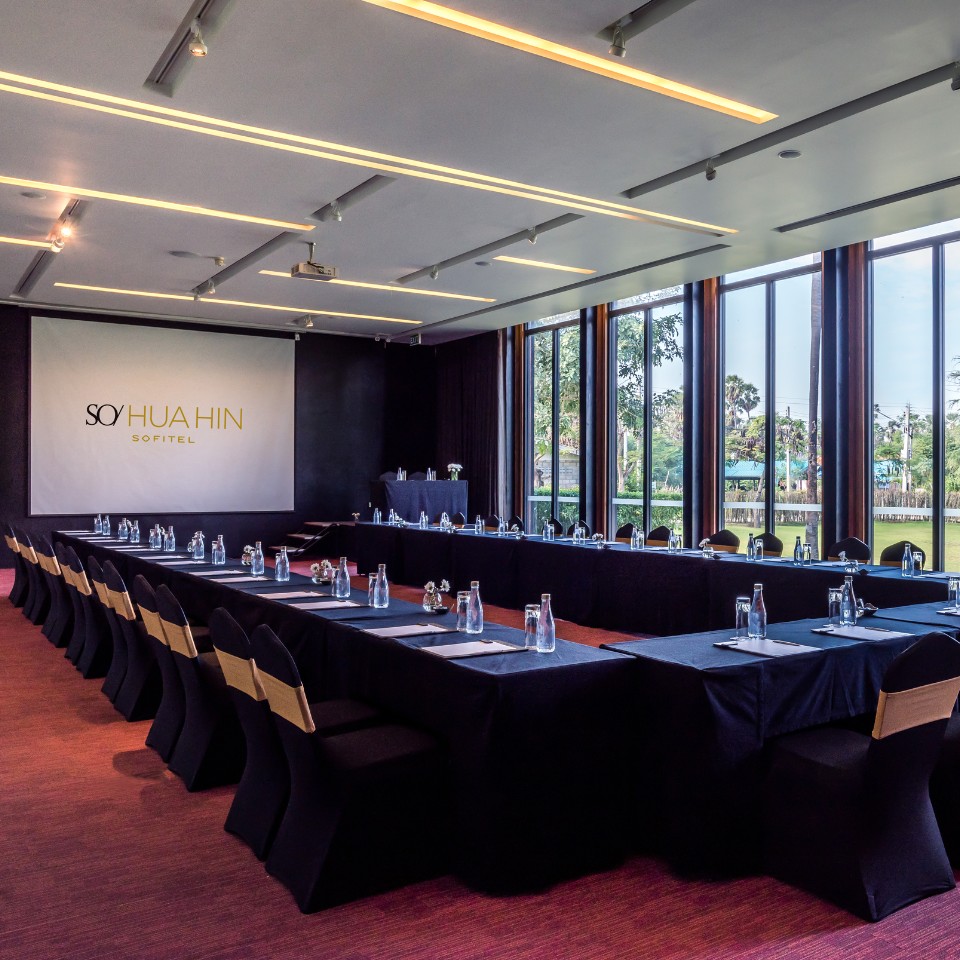 A conference room with a U-shaped table setup, black tablecloths and chairs, water bottles, and notepads. Large windows offer views of greenery outside.