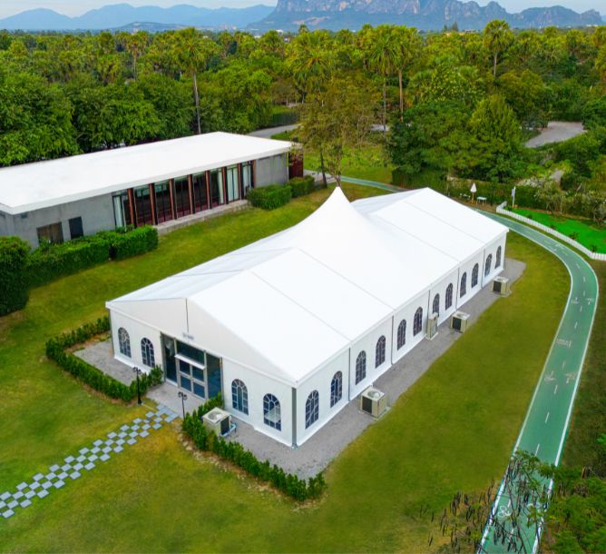 A white tent-like building with arched windows sits on a lush green lawn, surrounded by trees. A track curves around it, with mountains in the background.