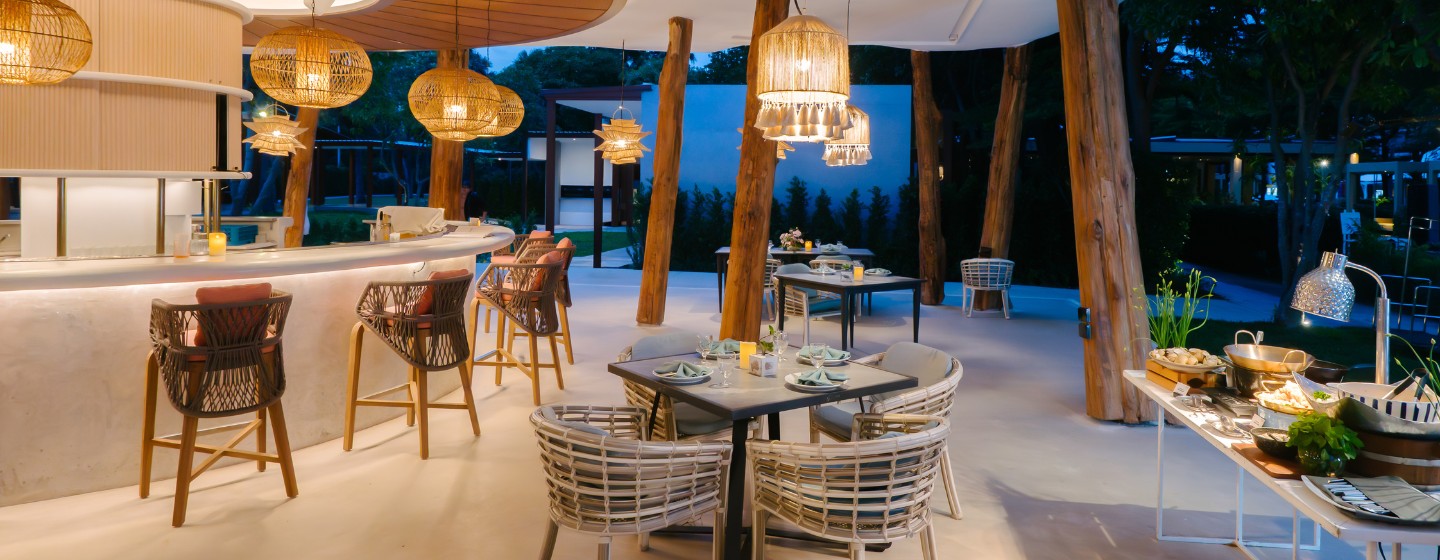 Stylish outdoor restaurant with wicker chairs, wooden tables, and warm hanging lights. The open-air setting conveys a cozy, inviting atmosphere.