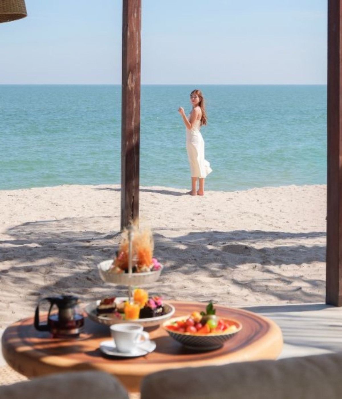 A serene beach scene features a woman in a white dress facing the ocean. In the foreground, a table holds a coffee set and fruit platter, conveying a tranquil, leisurely atmosphere.