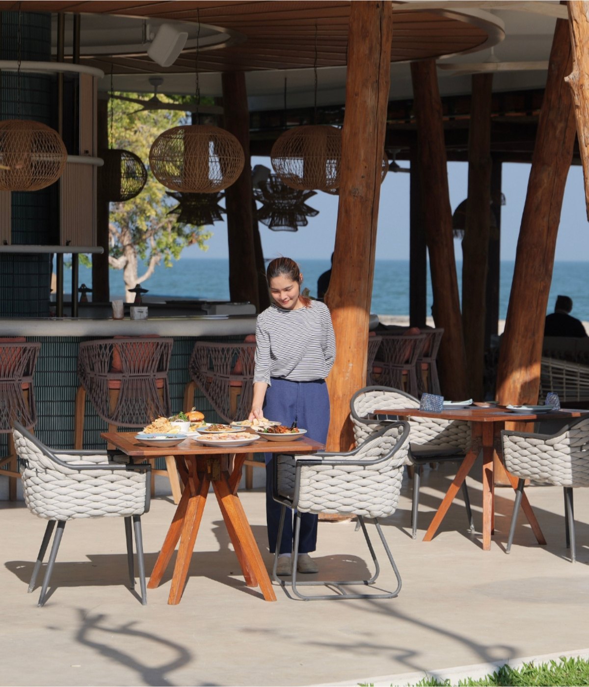 A woman in a striped shirt stands by a wooden table with dishes outdoors at a beachside restaurant. Wicker chairs and ocean view enhance the serene ambiance.