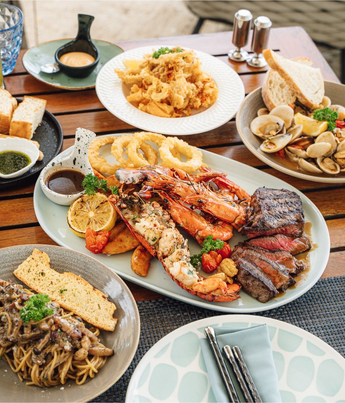 A wooden table displays a seafood and steak feast: grilled lobster, shrimp, and steak on a platter, surrounded by clam pasta, fries, and sauces. Cozy and inviting.