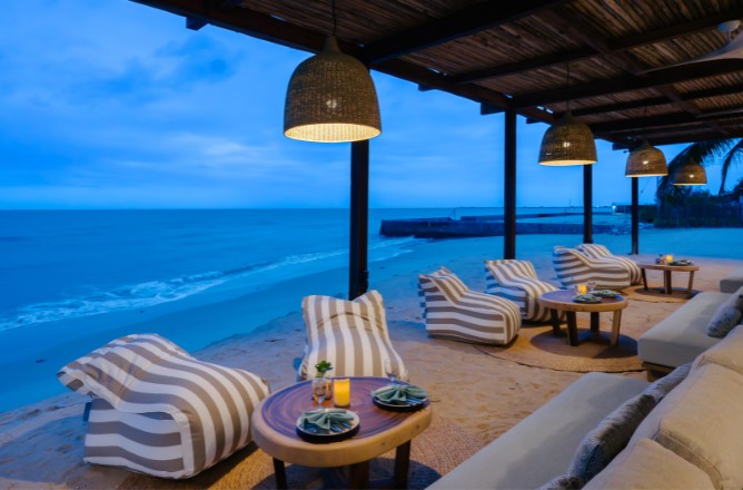 http://Outdoor%20beachside%20lounge%20at%20dusk%20with%20striped%20cushioned%20chairs,%20round%20tables,%20and%20warm%20pendant%20lights%20under%20a%20wooden%20canopy,%20overlooking%20a%20calm%20sea.