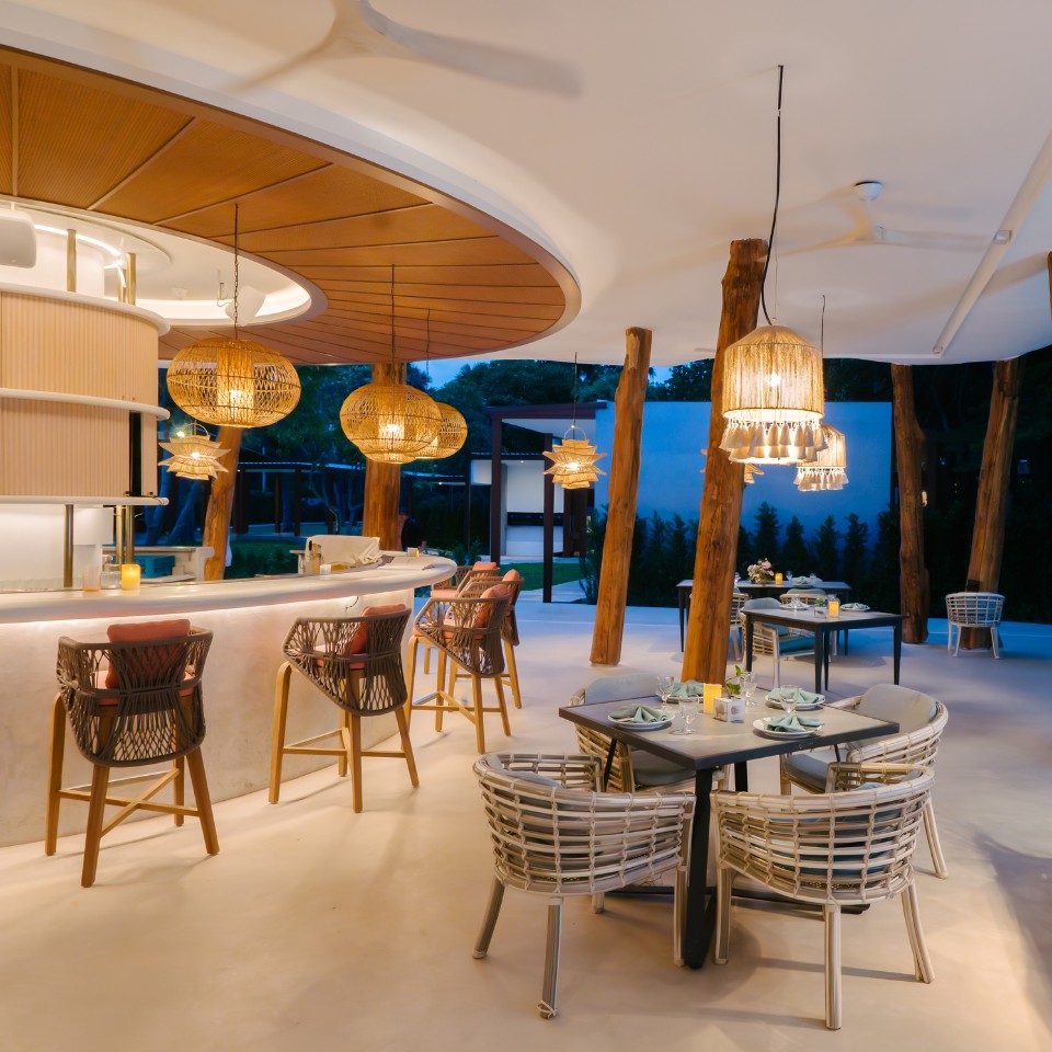 Stylish outdoor restaurant with wicker chairs, wooden tables, and warm hanging lights. The open-air setting conveys a cozy, inviting atmosphere.