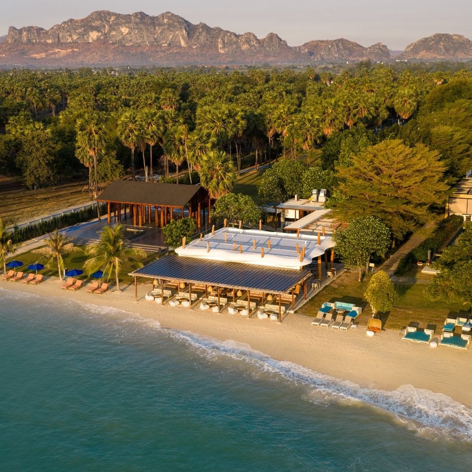 A serene beachfront scene with lush greenery, modern buildings, soft sand, and gentle waves at dusk. The atmosphere is tranquil and inviting.
