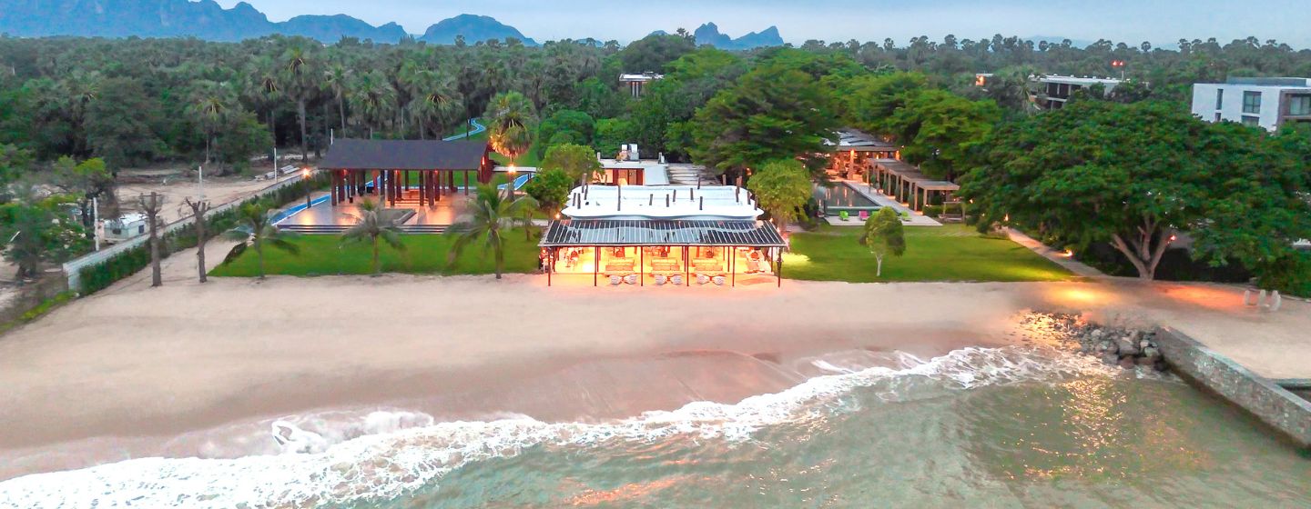 A serene beachfront scene with lush greenery, modern buildings, soft sand, and gentle waves at dusk. The atmosphere is tranquil and inviting.