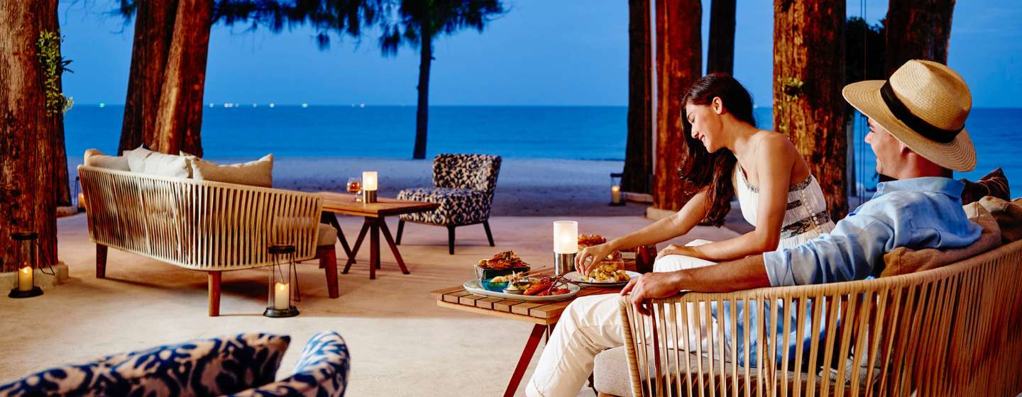 A couple relaxes on a patio with modern furniture, enjoying a meal by candlelight. They overlook a serene beach and calm sea at twilight.