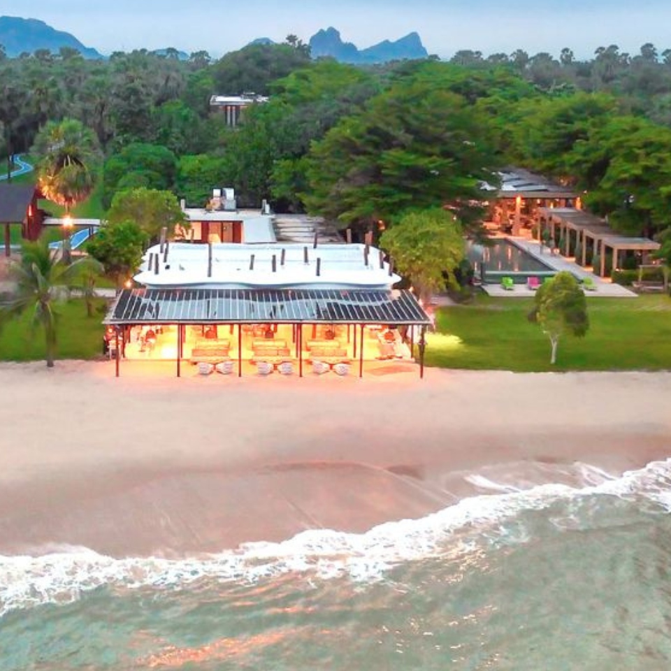 A serene beachfront scene with lush greenery, modern buildings, soft sand, and gentle waves at dusk. The atmosphere is tranquil and inviting.