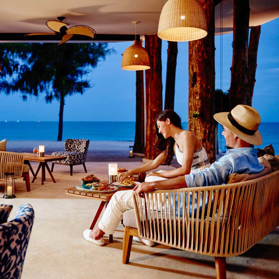 A couple relaxes on a patio with modern furniture, enjoying a meal by candlelight. They overlook a serene beach and calm sea at twilight.