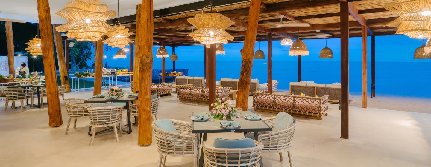 Beachfront restaurant with wicker furniture, wooden beams, and warm lighting. Overlooks a serene ocean at sunset, creating a cozy and inviting atmosphere.