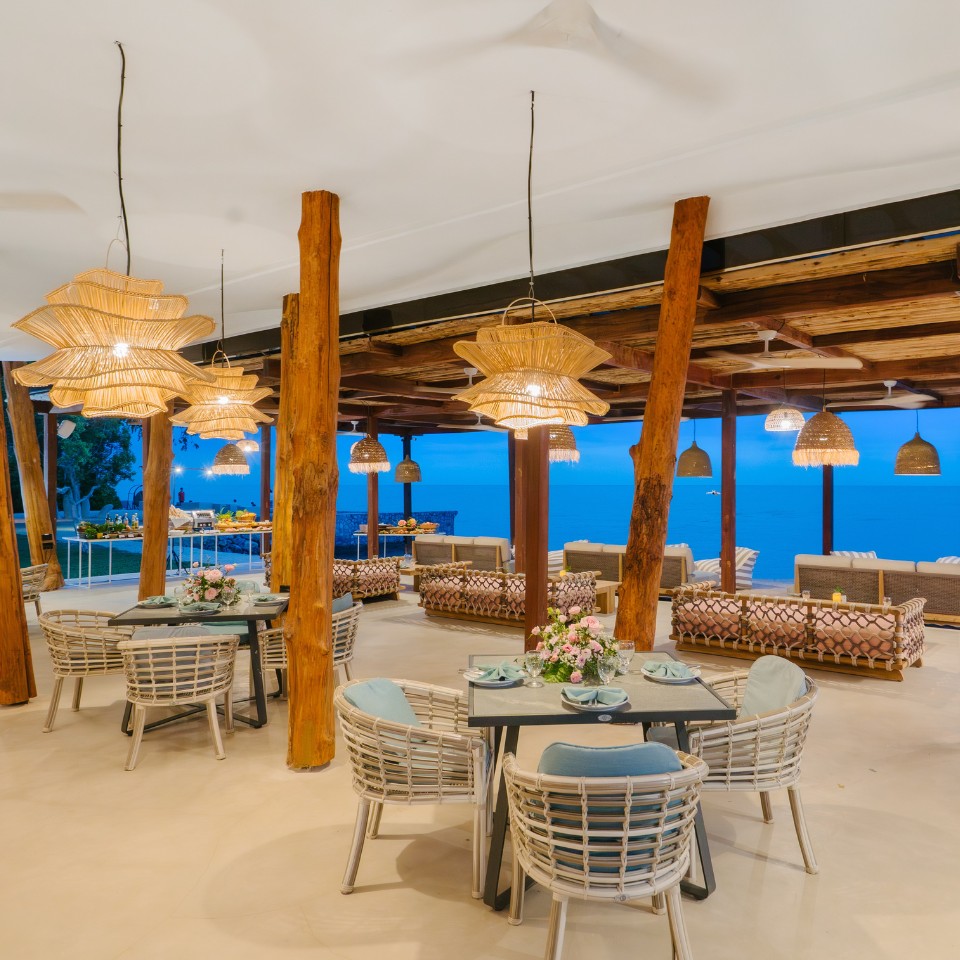 Beachfront restaurant with wicker furniture, wooden beams, and warm lighting. Overlooks a serene ocean at sunset, creating a cozy and inviting atmosphere.