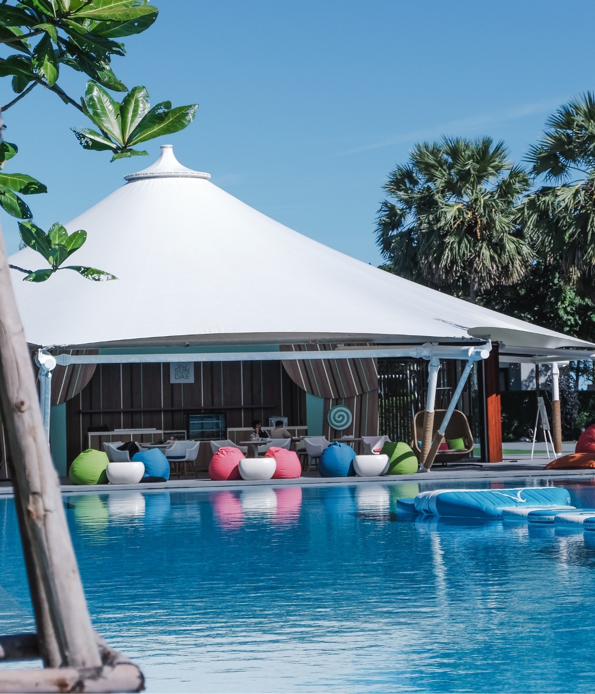 A serene pool scene with a large white gazebo and colorful bean bags under a clear blue sky. Palm trees and lush greenery surround the tranquil setting.