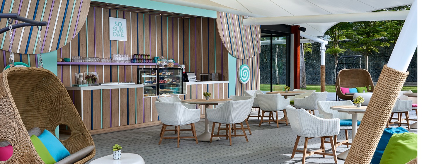 Outdoor patio with wicker furniture and colorful cushions, featuring a striped wall with a "So Sundae" sign, creating a tropical, relaxed vibe.