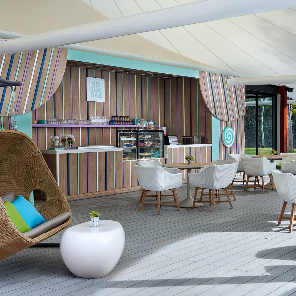 Outdoor patio with wicker furniture and colorful cushions, featuring a striped wall with a "So Sundae" sign, creating a tropical, relaxed vibe.