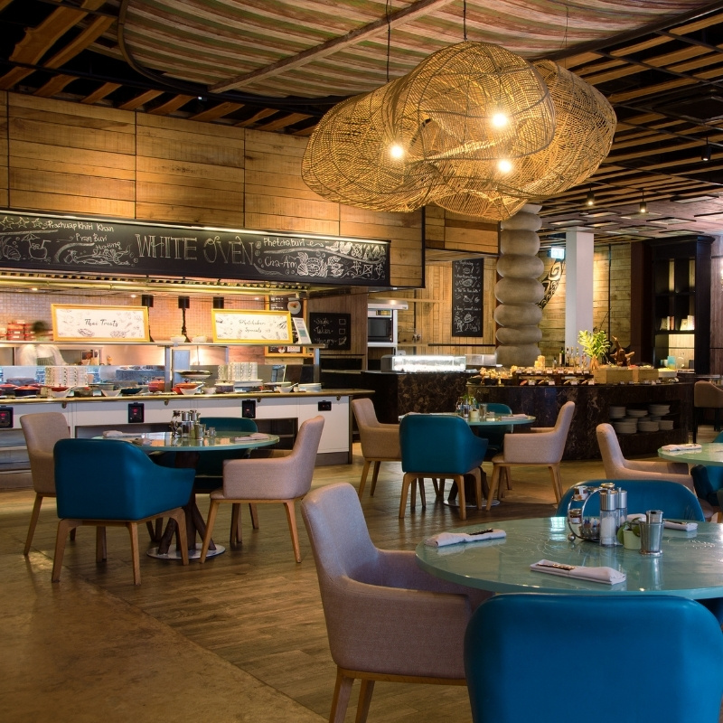 Modern restaurant interior featuring a mix of blue and beige chairs around a turquoise table, colorful bar stools, and woven light fixtures. Warm and inviting.