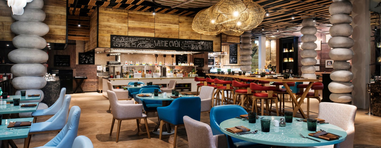 Modern restaurant interior with a warm, inviting ambiance. Features stacked stone columns, turquoise tables, beige and red chairs, and woven light fixtures.