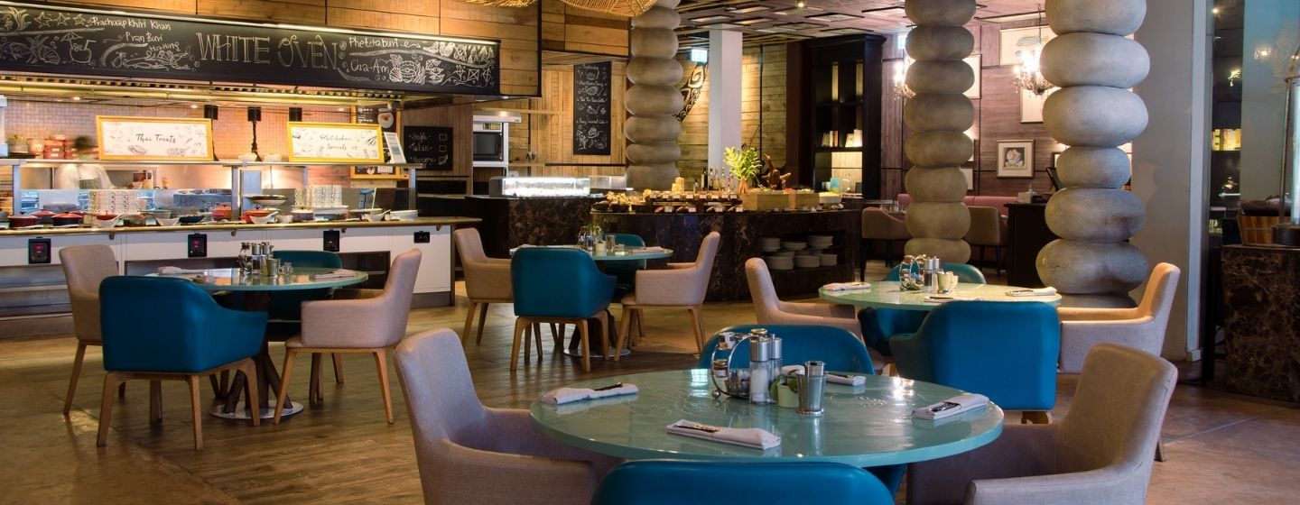 Modern restaurant interior with a warm, inviting ambiance. Features stacked stone columns, turquoise tables, beige and red chairs, and woven light fixtures.